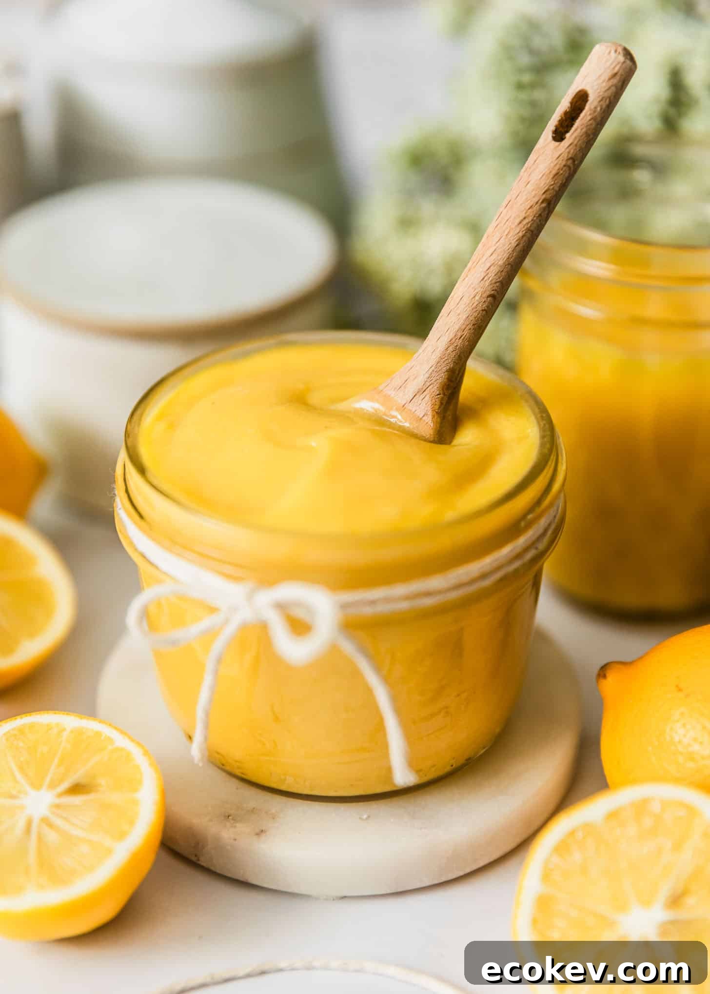 A jar of Meyer lemon curd with a wood spoon on a white counter next to lemons, white bowls, and green flowers.