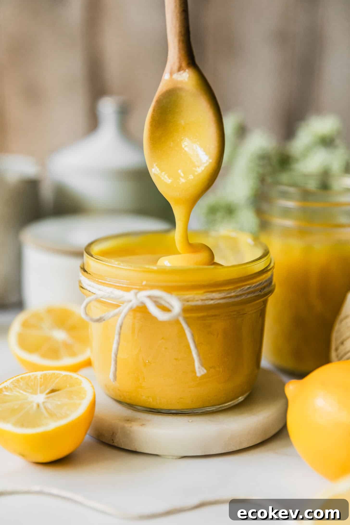 A spoon drizzling Meyer lemon curd in a jar on a white counter next to white bowls, green flowers, and lemons.
