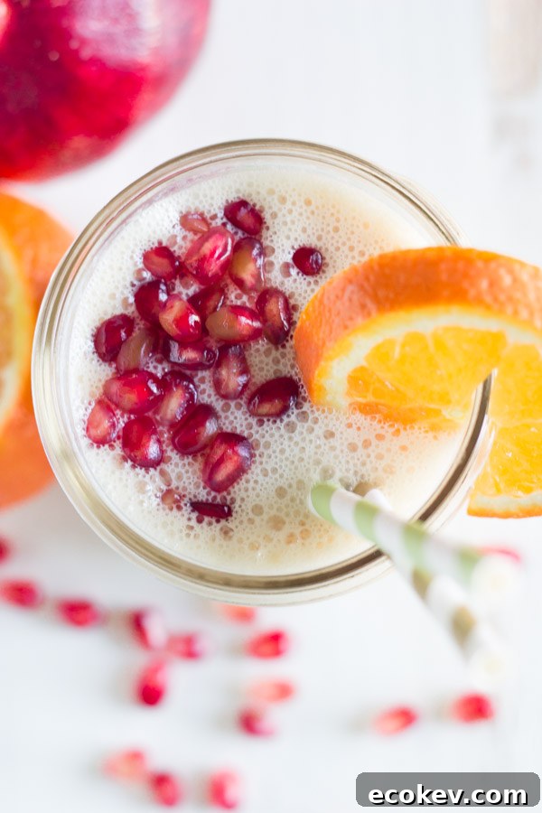 Pomegranate Citrus Smoothie in a glass jar with pomegranate seeds on top, an orange slice on the rim of the glass and two straws in it.