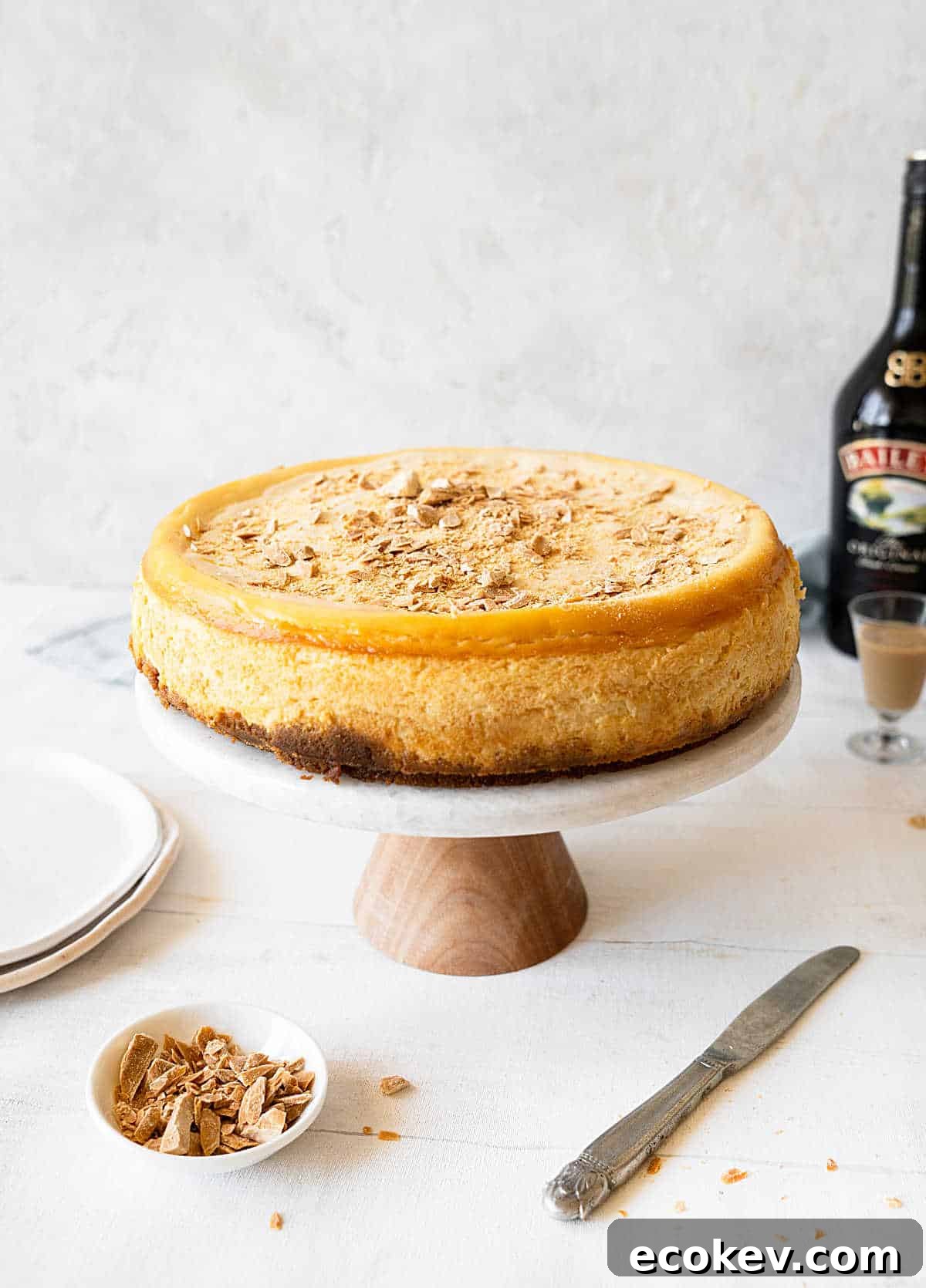 Whole Baileys cheesecake on a cake stand. Light gray background and white surface. White plates, knife, bottle. 