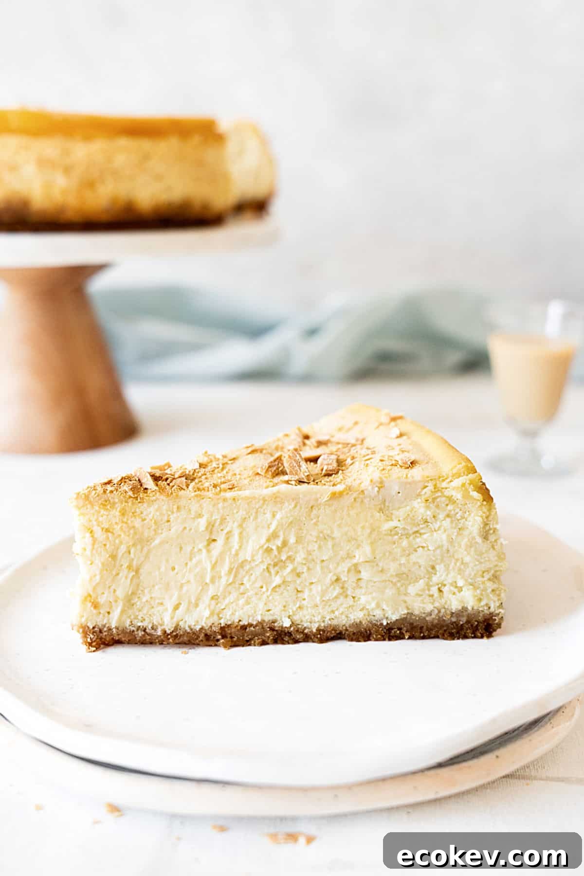 Slice of Baileys cheesecake on a white plate. Light gray background with cake stand, light blue cloth. 
