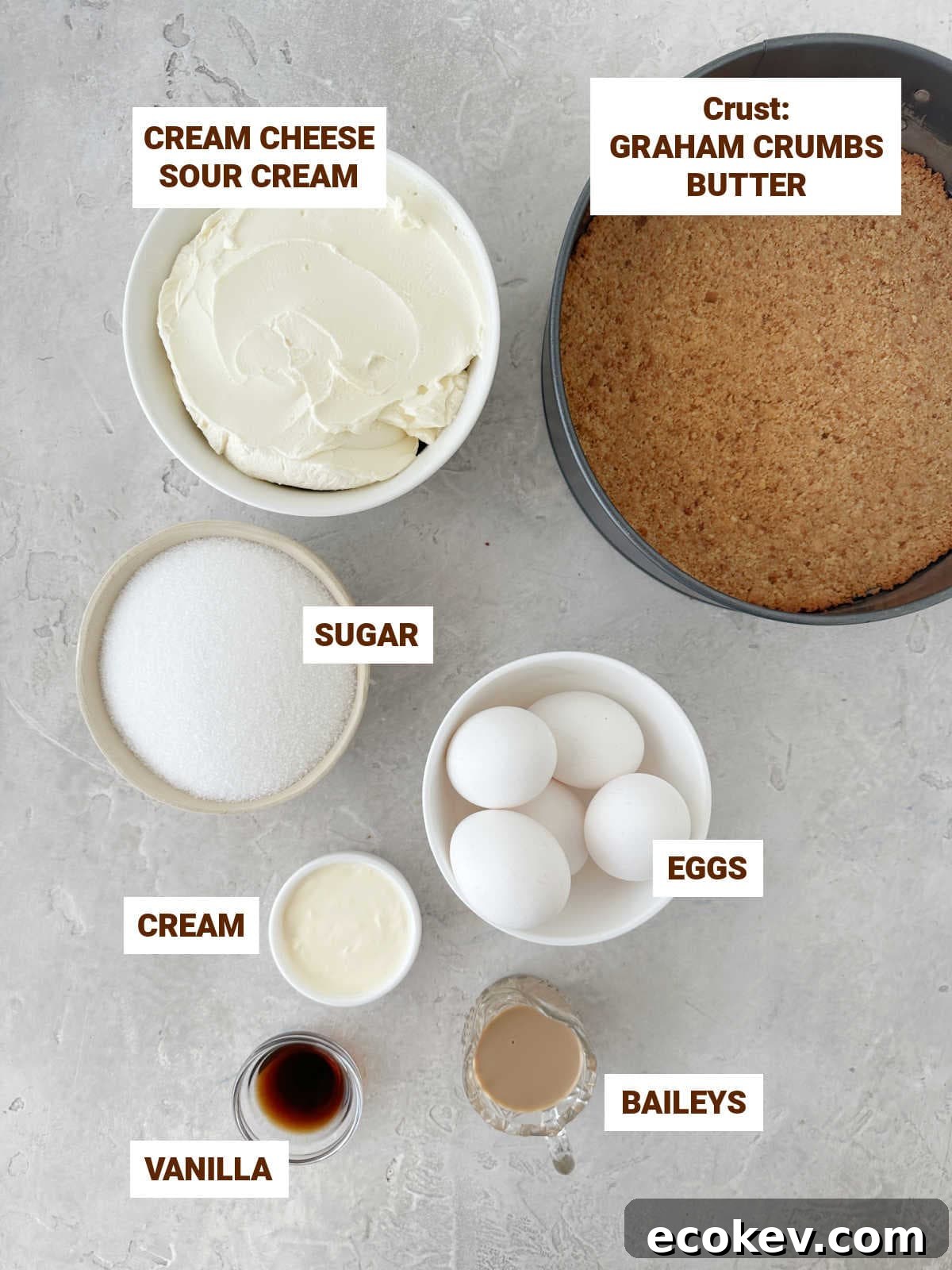 Gray surface with bowls containing ingredients for Baileys cheesecake including graham base, eggs, vanilla, sugar, cream.