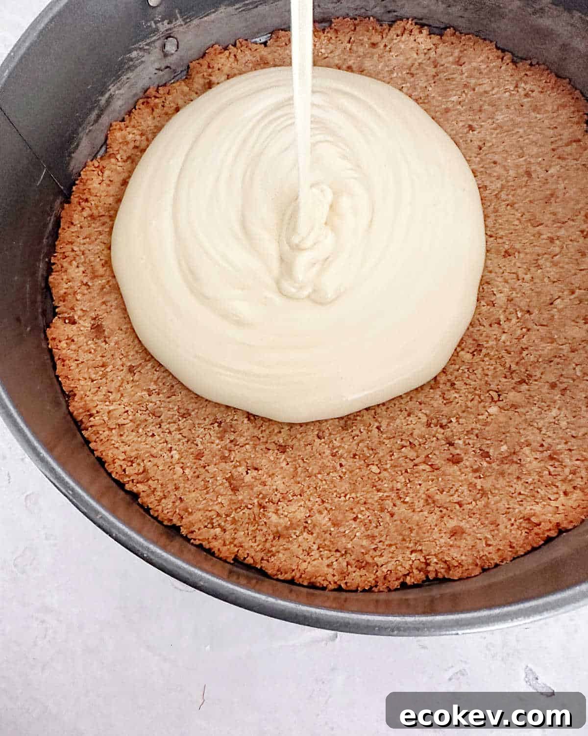 Cheesecake batter being poured on a graham crust in a metal pan. Light grey surface.