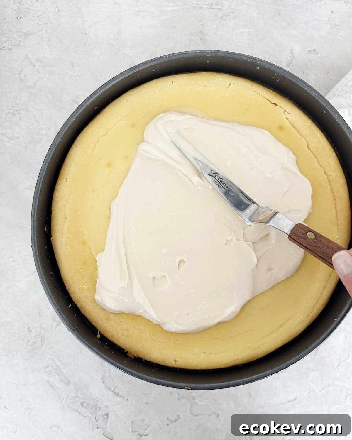 Spreading sour cream topping on a baked cheesecake in a metal pan. Light grey surface. 