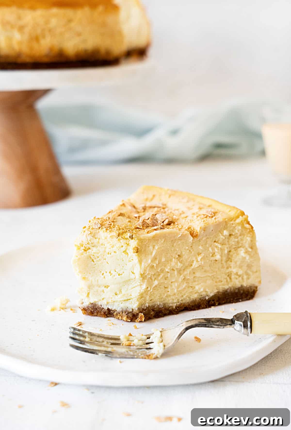 Bitten slice of Baileys cheesecake on a white plate. Silver fork. Cake stand and light blue cloth in the background. 