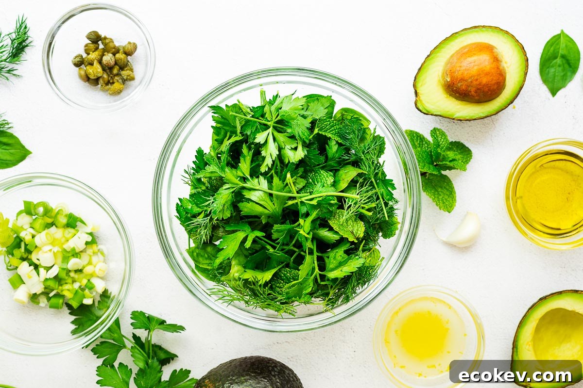 Chopped green herbs surrounded by the rest of the ingredients for vegan green goddess dressing, including sliced avocado, olive oil, lemon juice and zest, chopped green onions and capers.