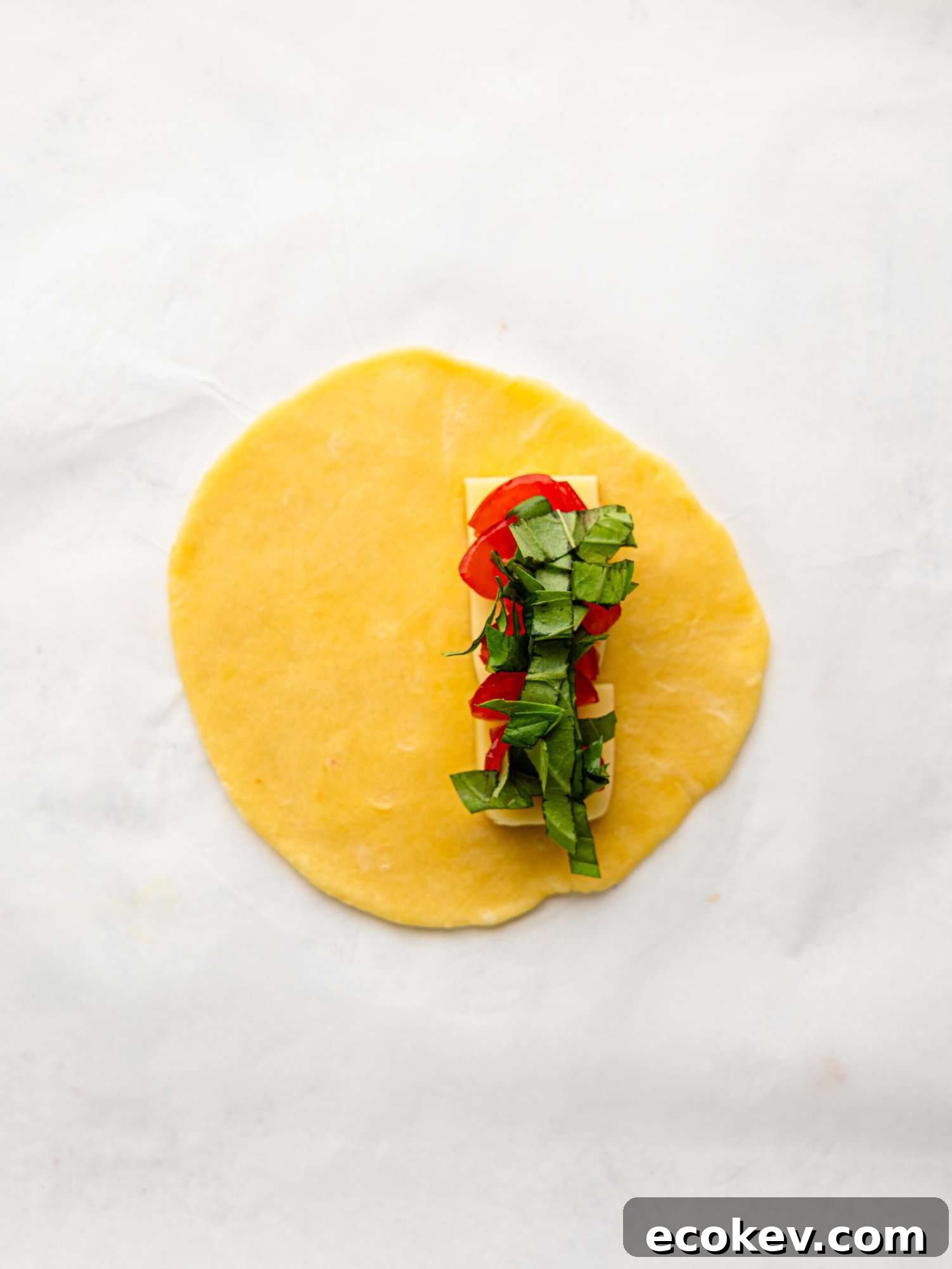 An empanada disk with mozzarella, tomatoes and basil on a sheet of parchment paper.