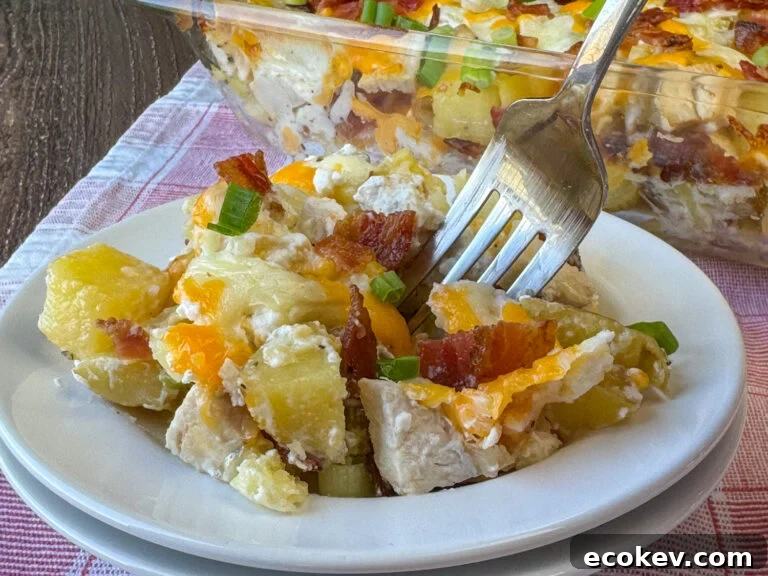 Close up of a fork collecting a bite of small cubes of chicken and potatoes with cheese, sour cream and bacon on a white plate with the full pan in the background.