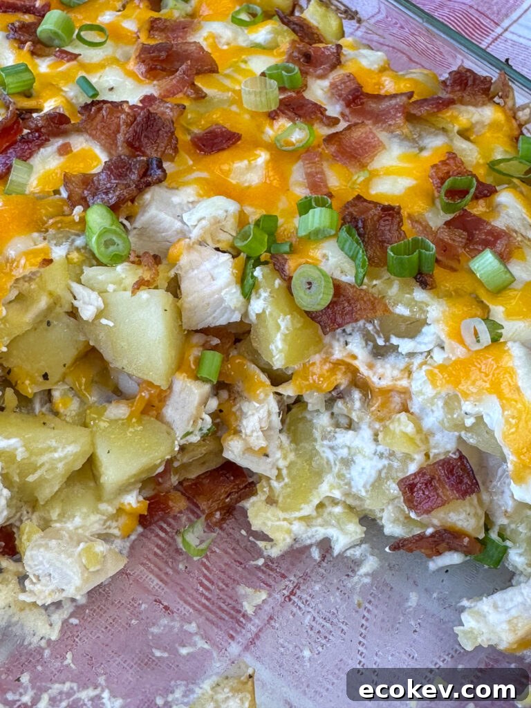 Close up layer of melted cheese oozing over top tender cubes of chicken and potatoes that are nestled in sour cream and generous pieces of bacon and accented with slices of green onions in a glass casserole dish.