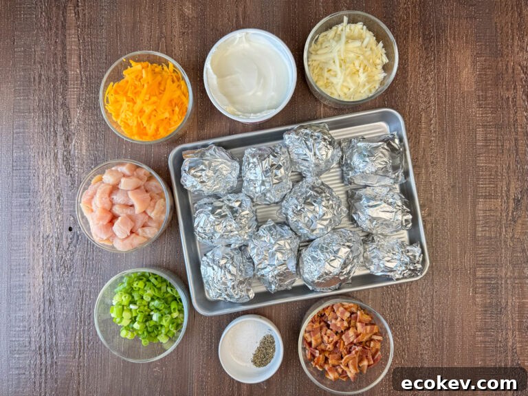 Overhead view of baked potatoes wrapped in foil surrounded by Monterey Jack cheese, sour cream, cheddar cheese, cubes of chicken breast, green onions, salt, pepper and chopped bacon on a wooden counter top.