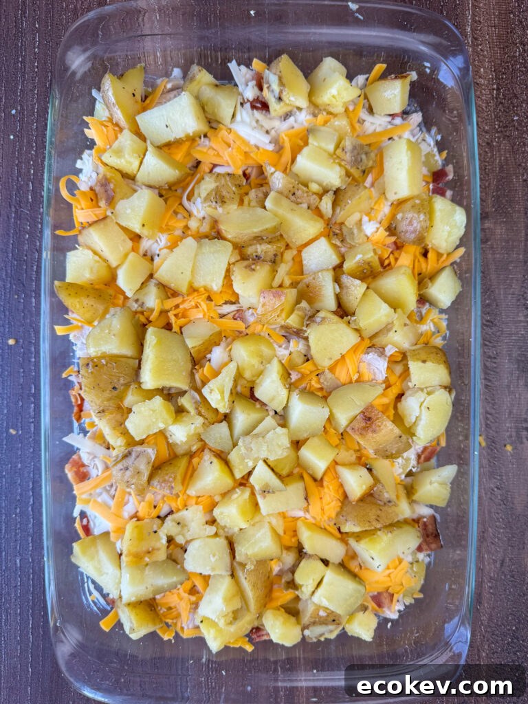 Second layer of chopped baked potatoes spread over a cheesy and sour cream mixture that's separating layers of chicken and potatoes in a clear glass 9 x 13