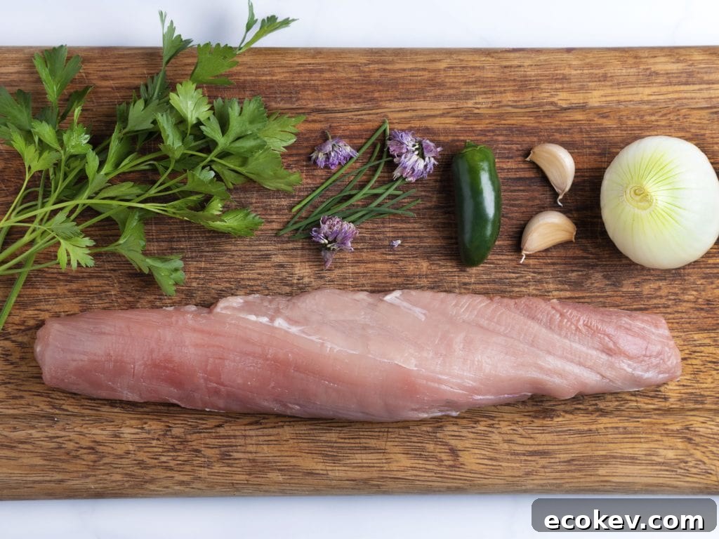 Ingredients on a cutting board: raw pork tenderloin, jalapeno pepper, fresh herbs and garlic