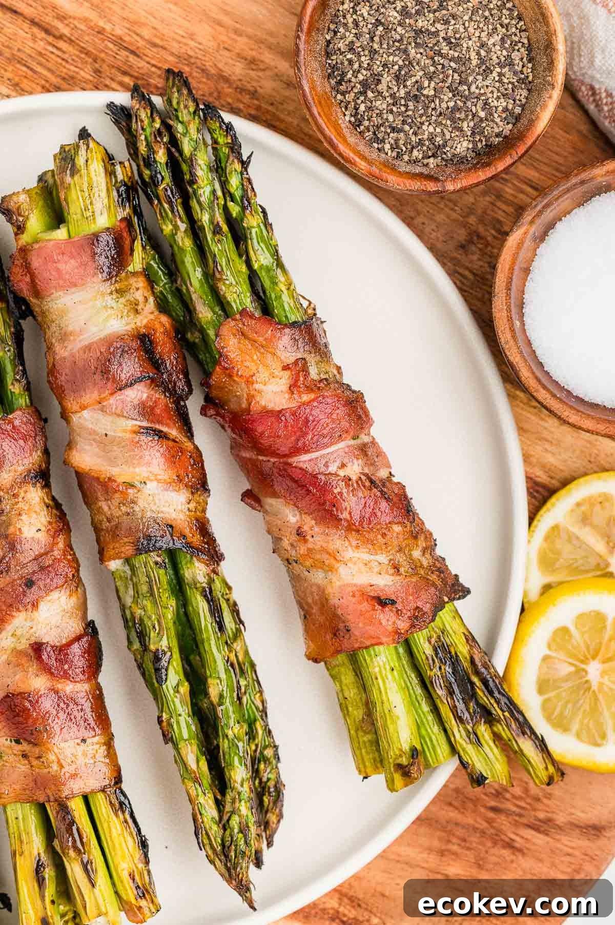 Two bundles of grilled asparagus wrapped in bacon on a white plate, with small bowls of pepper and salt, and lemon slices on the side.