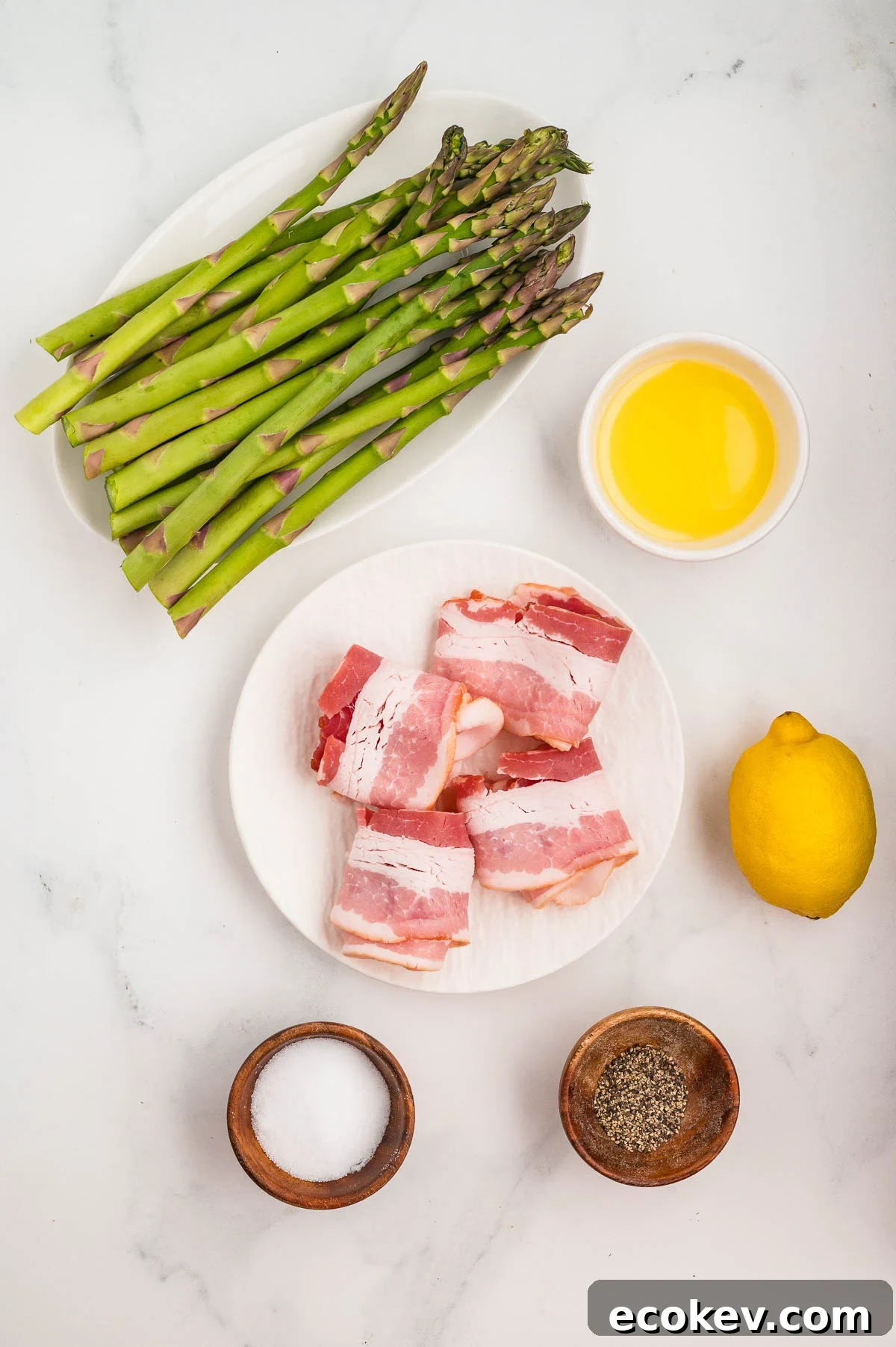 Asparagus, bacon-wrapped items on a plate, melted butter in a bowl, a whole lemon, and small bowls of salt and pepper on a white surface.