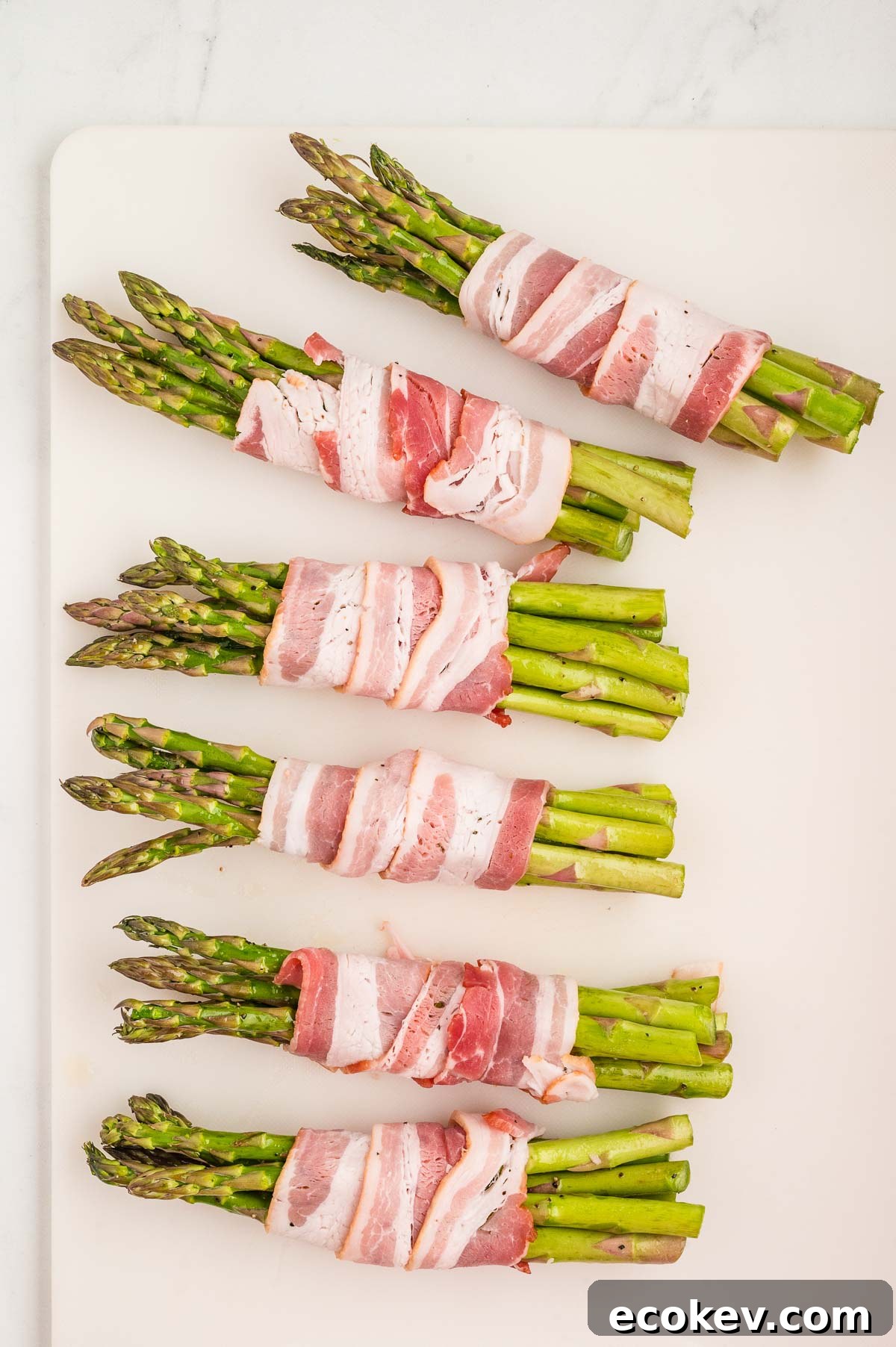 Six bundles of asparagus wrapped with bacon are arranged on a white surface.