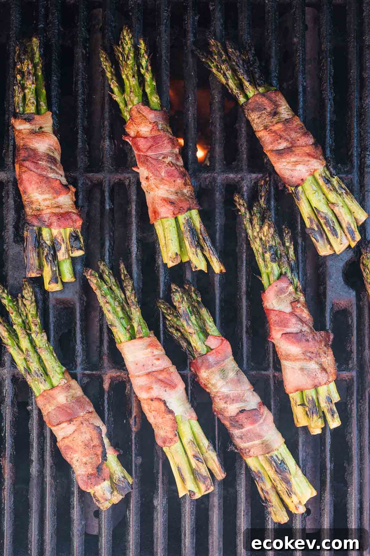 Asparagus bundles wrapped in bacon are grilling on a barbecue, displaying a mix of charred and crisp textures.