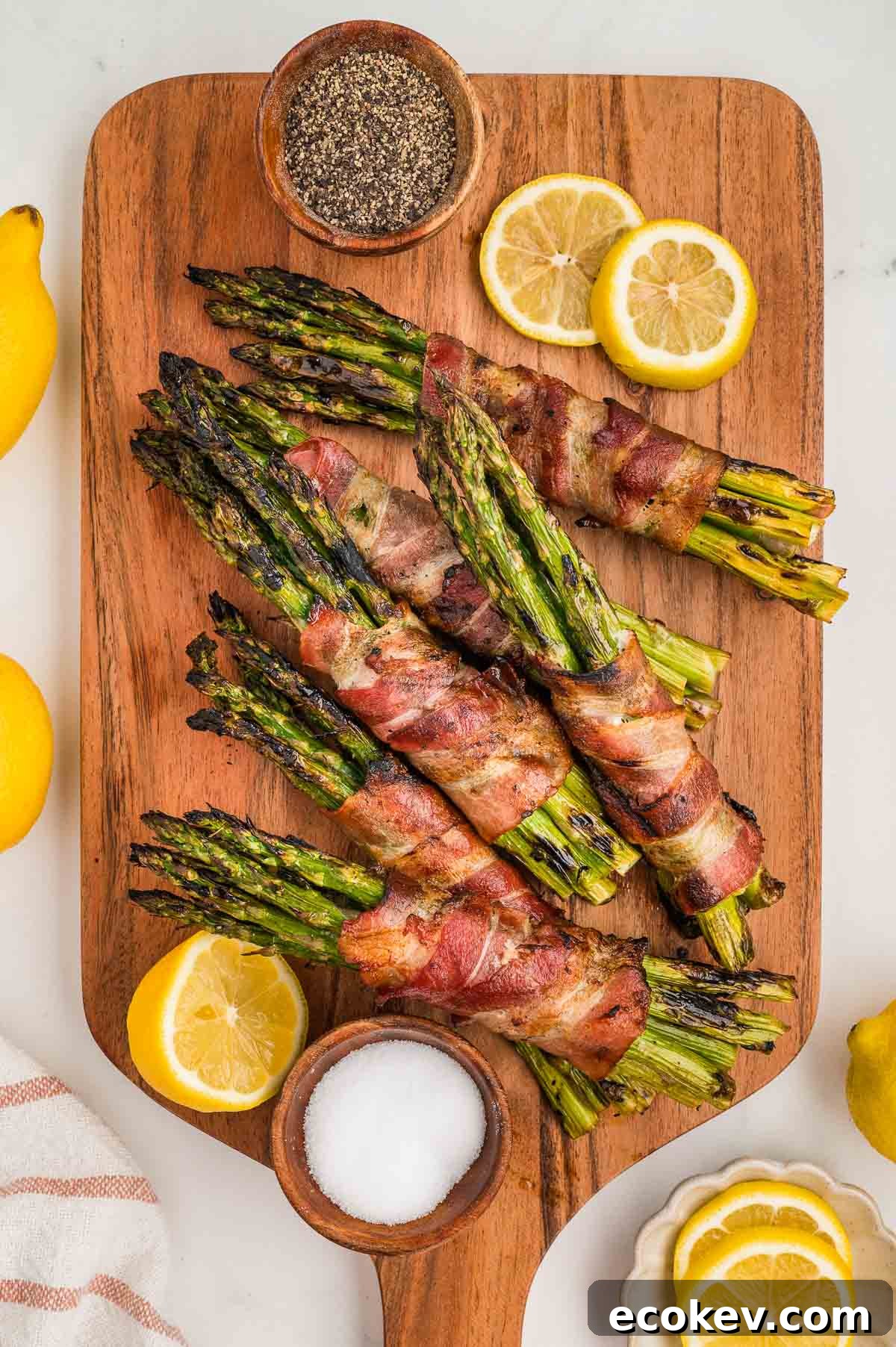 Bacon-wrapped asparagus bundles on a wooden board with bowls of black pepper and salt, garnished with lemon slices and whole lemons.