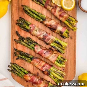 Bacon-wrapped asparagus bundles on a wooden board, garnished with lemon slices and bowls of salt and pepper.