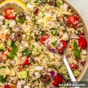 A close-up of a bowl of couscous salad with cherry tomatoes, cucumber, olives, red onion, feta cheese, and herbs.