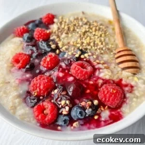 A bowl of oatmeal topped with fresh raspberries, blueberries, a honey drizzle, and a sprinkle of seeds, served with a wooden honey dipper on a light wooden table.