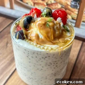 A glass of chia pudding topped with berries, blueberries, peanut butter, honey, and fresh herbs, set on a wooden table with pastries visible in the background.