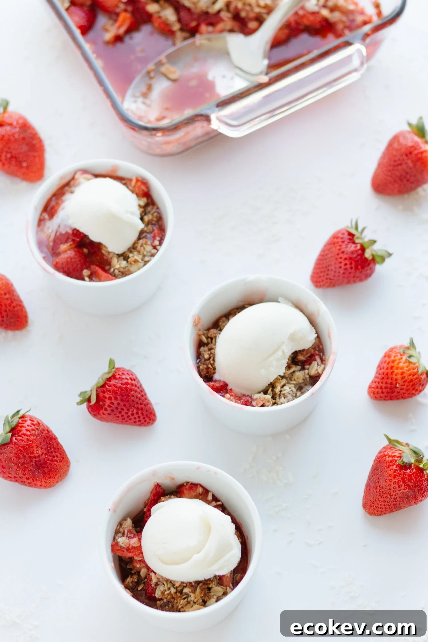 Three ramekins of strawberry coconut crisp topped with ice cream  with baking dish in background and strawberries scattered on white surface.