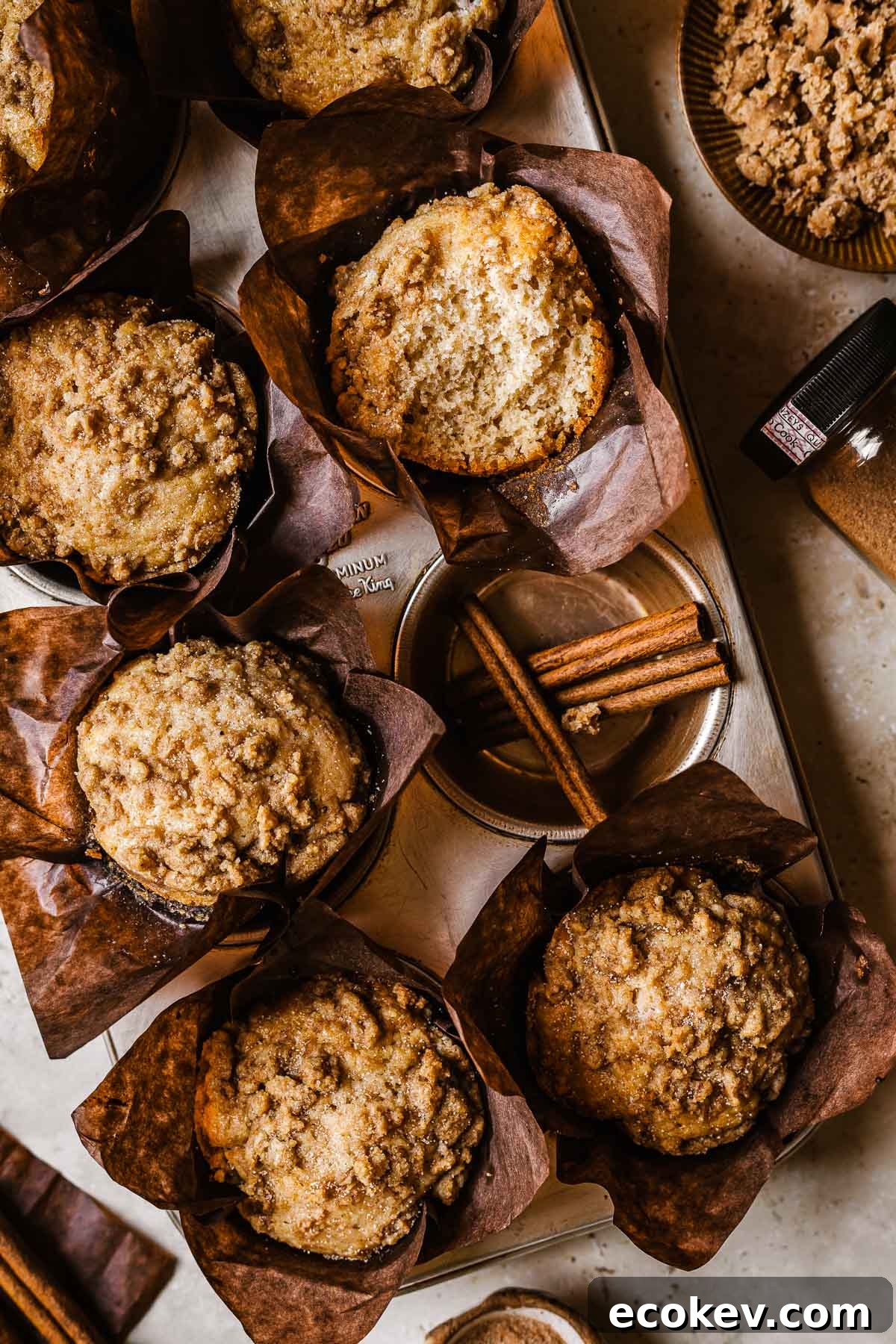 Cinnamon streusel muffins in brown parchment paper wrappers. One muffin has been broken open to show the crumb inside. 