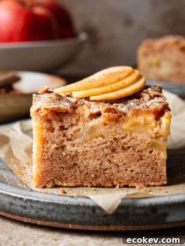 A slice of moist apple cake on a ceramic plate with three thin slices of fresh apple on top.