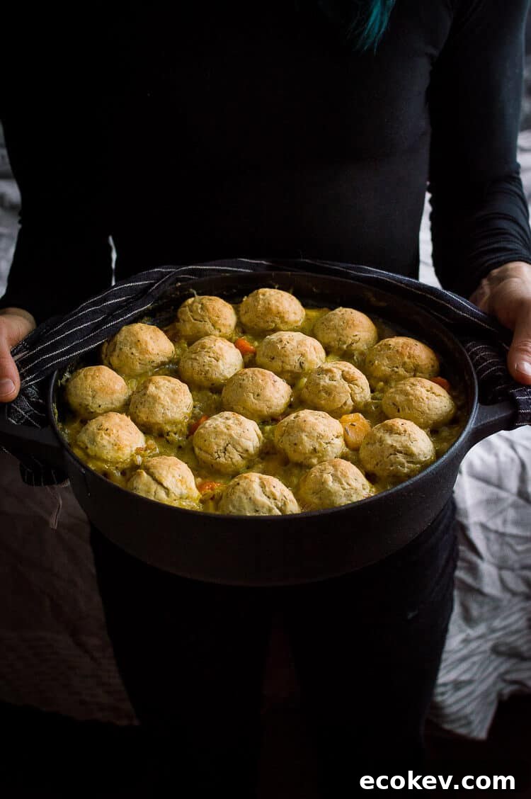 A black cast iron pan of vegan vegetable and pearl barley stew topped with herby dumplings being held by a person dressed in black.