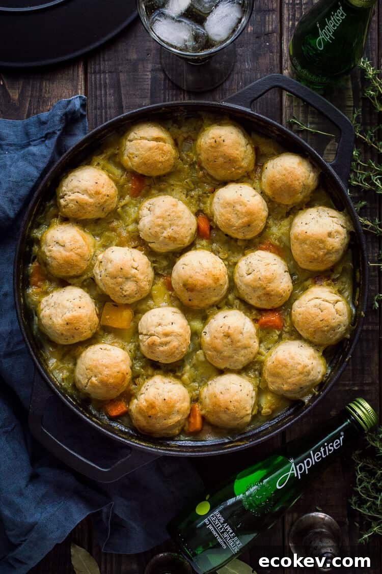 A cast iron pan of vegan vegetable and pearl barley stew with herby dumplings and a bottle of Appletiser on a dark wood background.