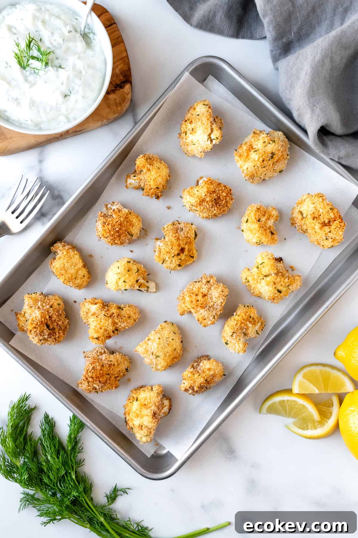 A parchment lined baking sheet with Crispy Cauliflower Bites on a marble countertop with a bowl of dill dipping sauce.