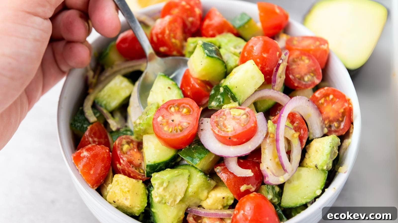 A hand holds a spoon in a bowl of avocado tomato salad with cucumber.