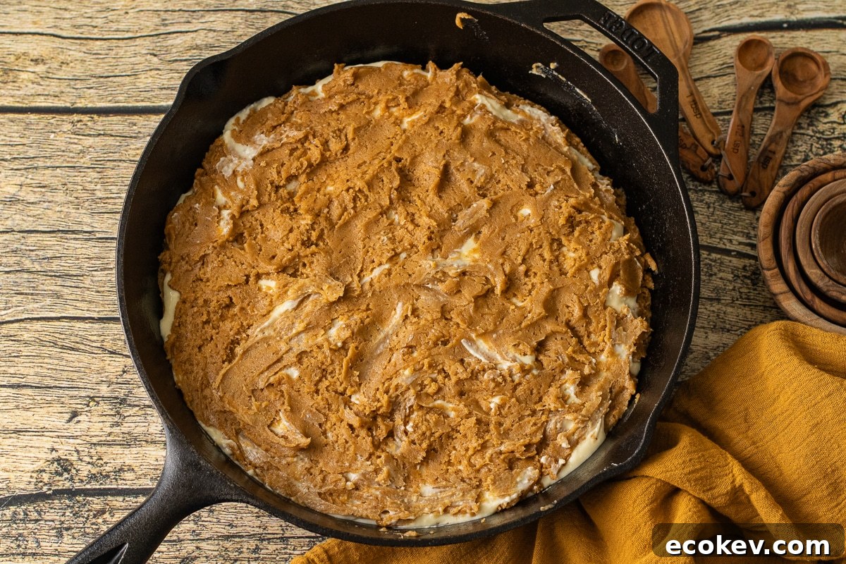 A cast iron skillet filled with a golden pumpkin cookie swirled with a creamy cheesecake layer uncooked