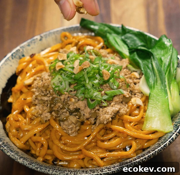 Authentic Dan Dan Noodles with Spicy Sichuan Sauce 7 Hand sprinkling crushed peanuts onto a bowl of dan dan noodles topped with pork, scallions, and bok choy.