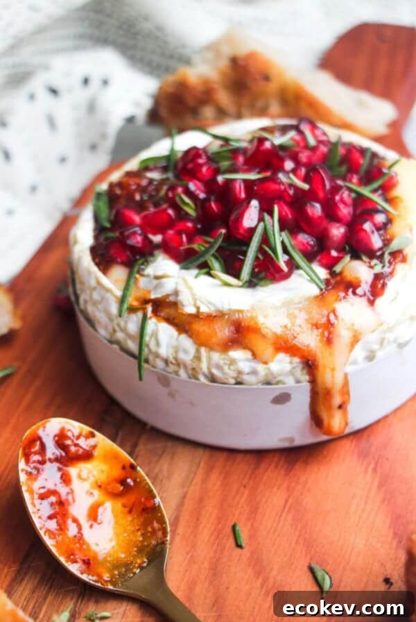 Wheel of camembert on a wooden board with oozy cheese spilling out, topped with pomegranate seeds and herbs.