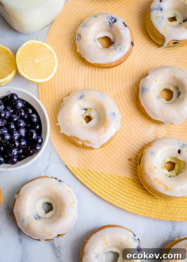 lemon blueberry donuts laid on a marble background with a yellow round place mat with a sliced lemon and blueberries beside the donuts