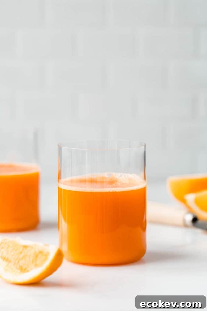 Two glasses or orange and carrot juice on a counter with sliced oranges around them