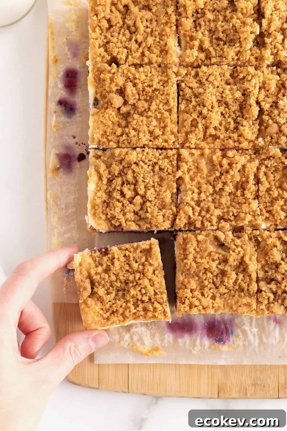 12 square lemon blueberry bars on a parchment lined cutting board. A hand is removing the bottom left bar using their thumb and forefinger.