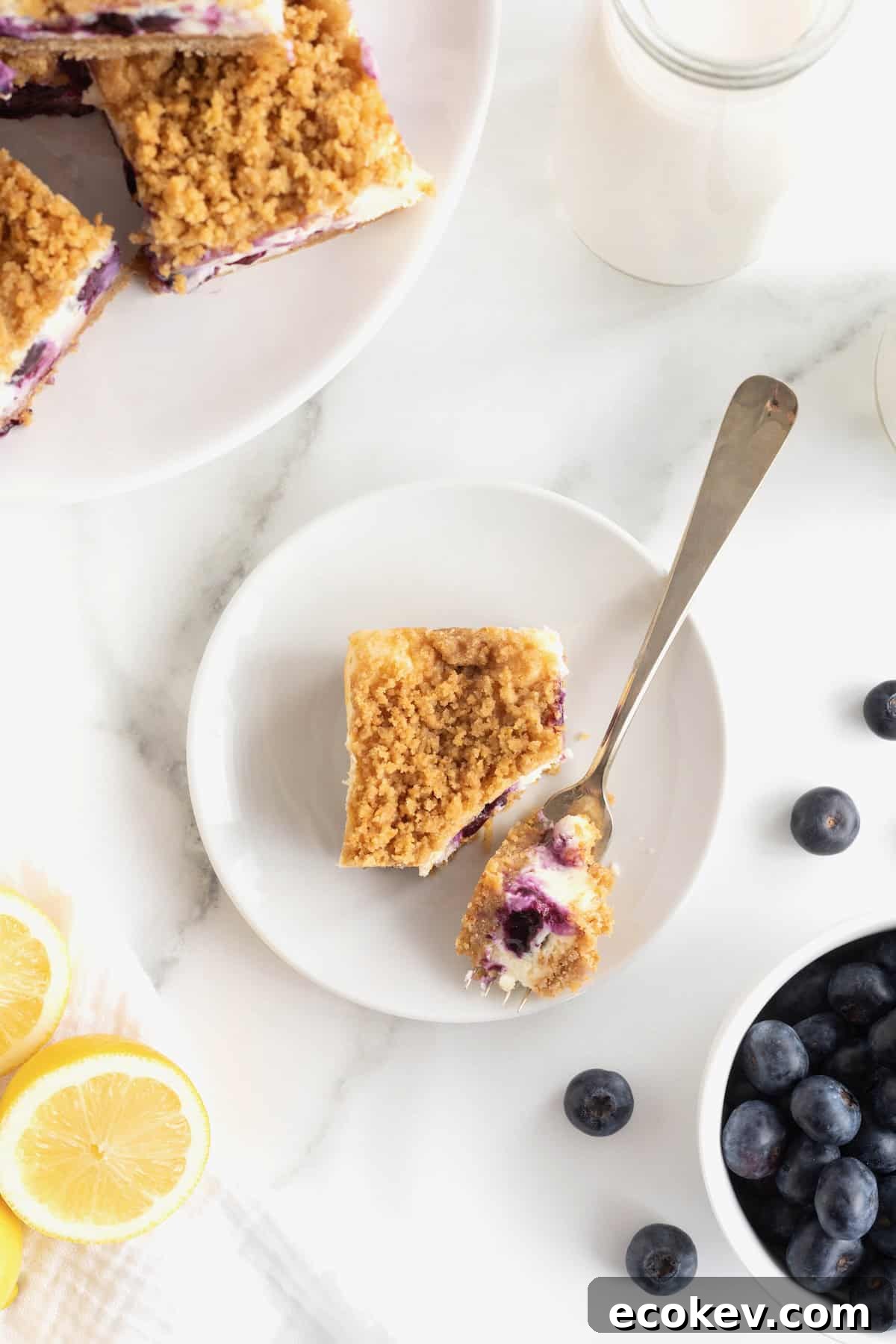A lemon blueberry cheesecake bar on a small white dessert plate. A fork rests on the plate with a bite of the dessert on it.