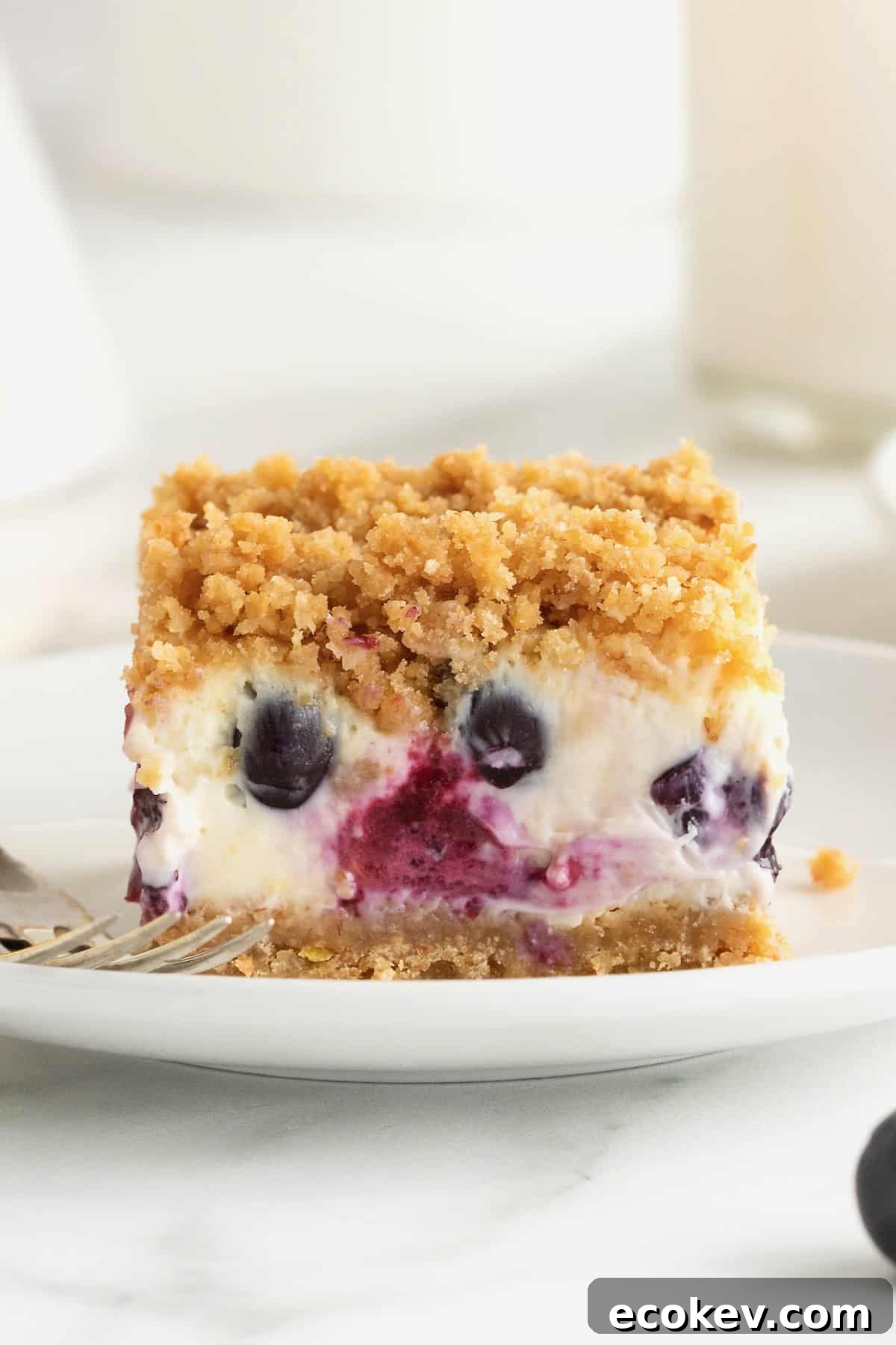 A side view of lemon blueberry cheesecake bars on a small white dessert plate. A fork rests to the left of the food.