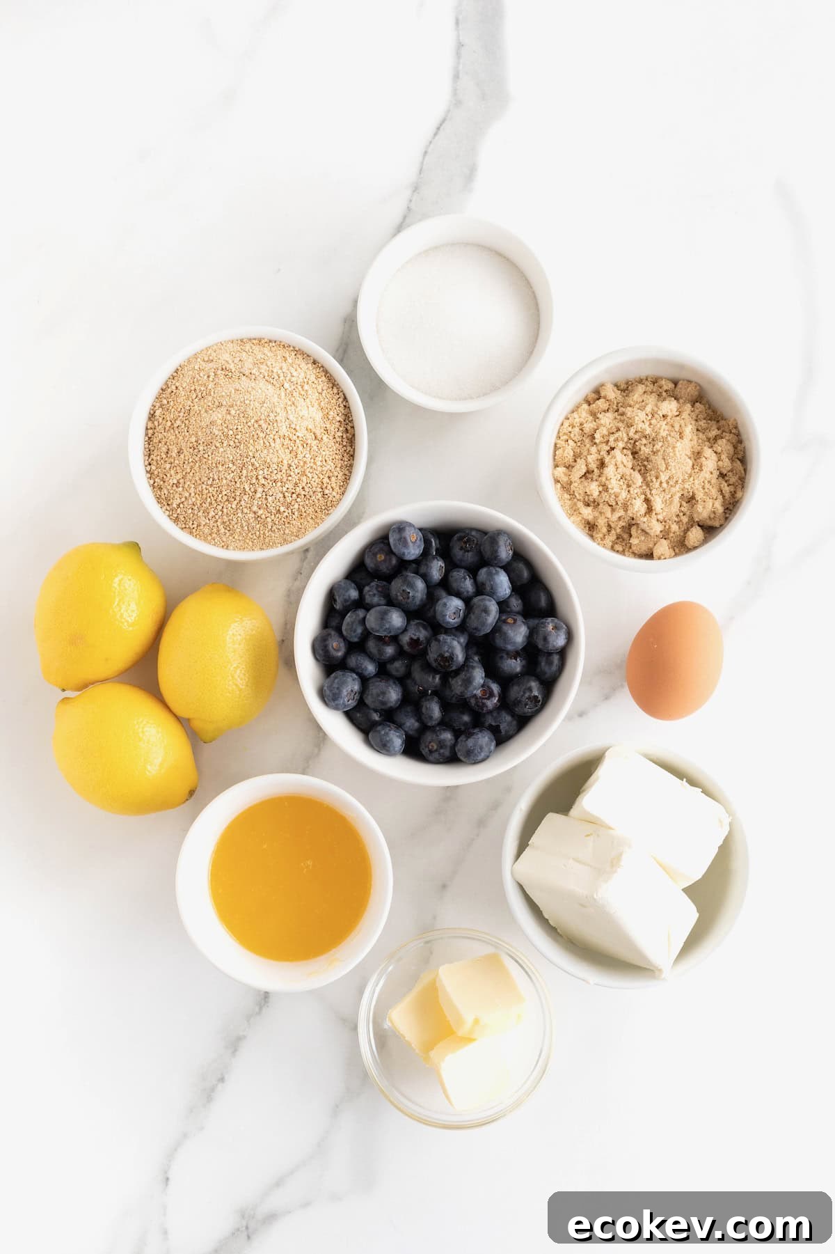 Ingredients to make lemon blueberry cheesecake bars in small white dishes on a white marble counter.
