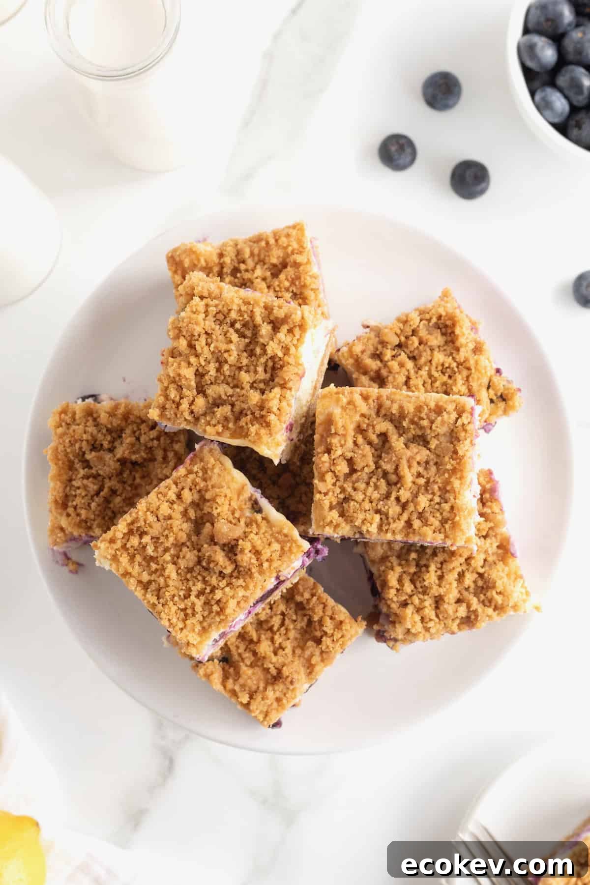 An overhead view of eight cheesecake bars on a large white serving plate. Blueberries are scattered at the top right edge of the plate.