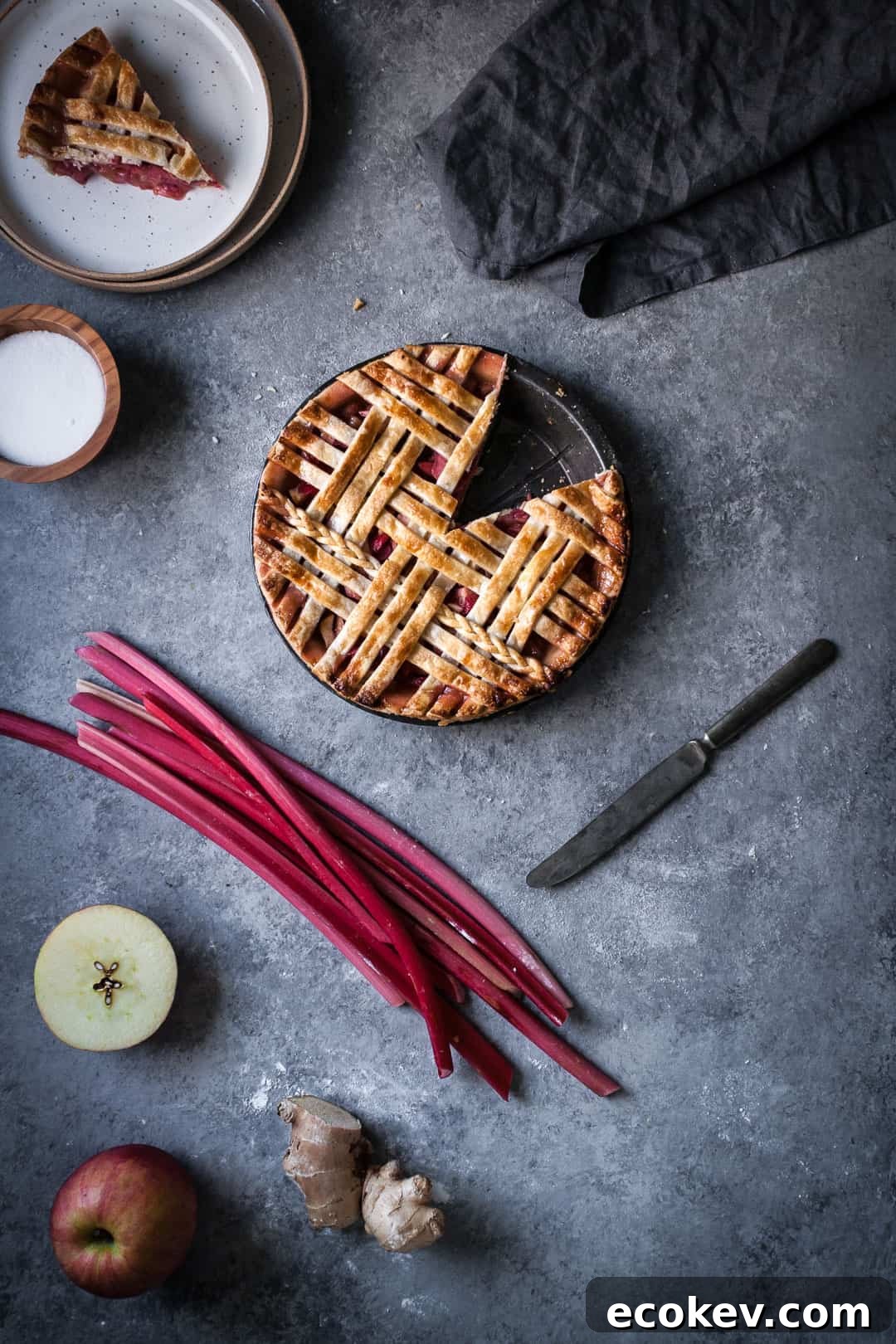 baked rhubarb apple pie with ginger and lemongrass on a grey surface with a piece cut out and the pie ingredients surrounding it