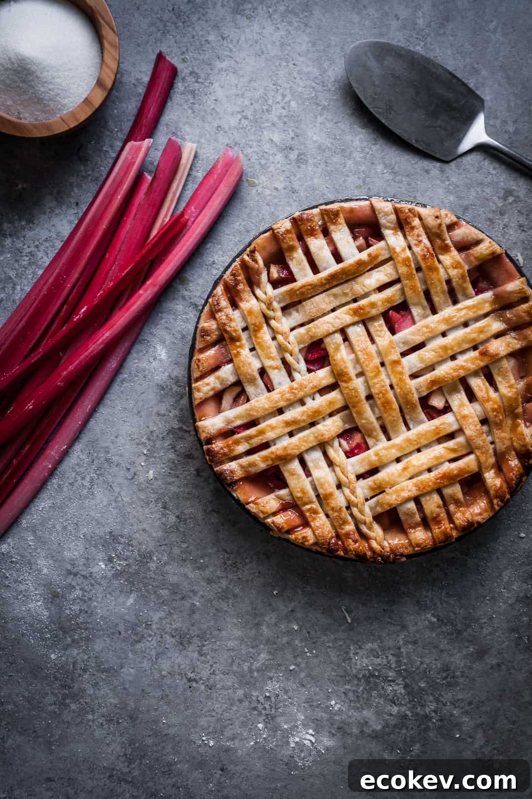 close up on grey background of baked rhubarb apple pie with ginger and lemongrass rhubarb stalks and pie server nearby