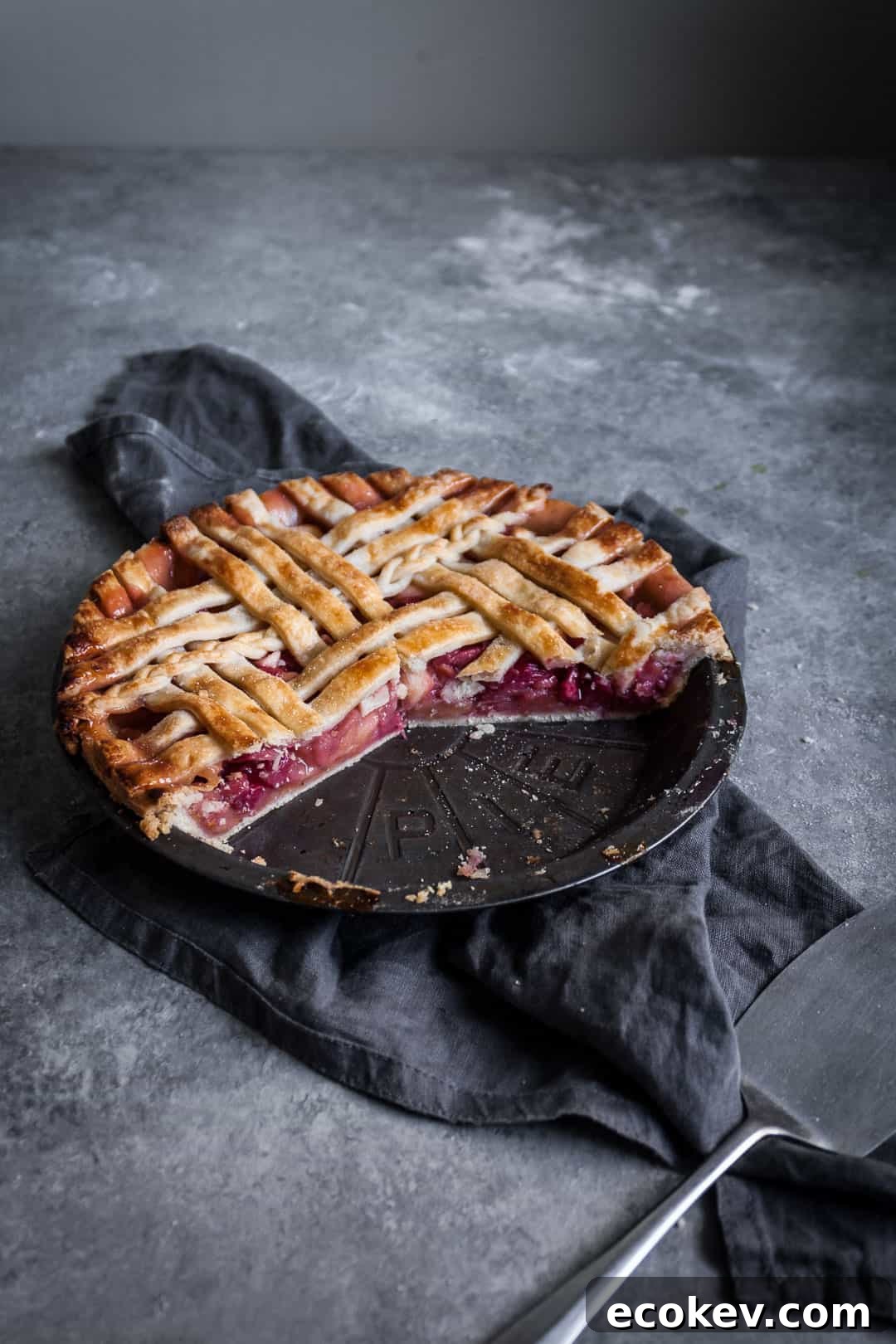 angled shot of rhubarb apple pie with ginger and lemongrass on a grey surface with a piece cut out on a plate and a grey linen towel underneath the pie tin