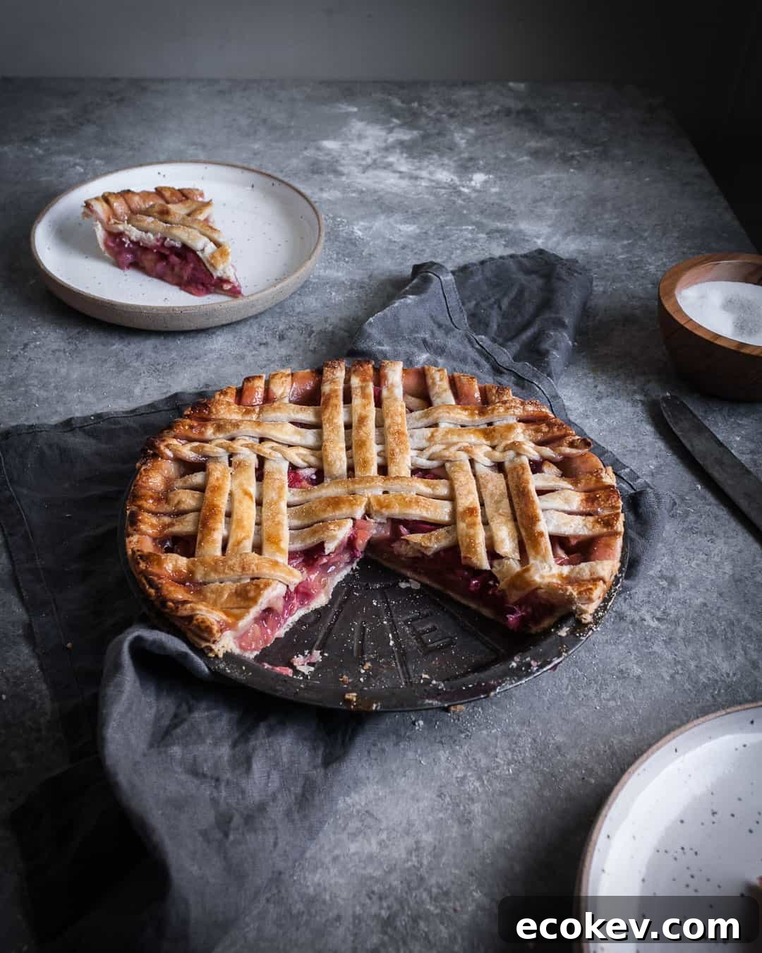 angled shot of rhubarb apple pie with ginger and lemongrass on a grey surface with a piece cut out on a plate and a grey linen towel underneath the pie tin