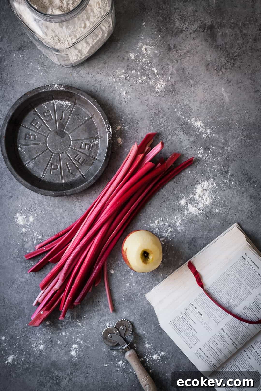making a rhubarb apple pie with ginger and lemongrass: top view of rhubarb stalks and peeled apple on grey surface with opened cookbook, metal pie tin and flour container nearby