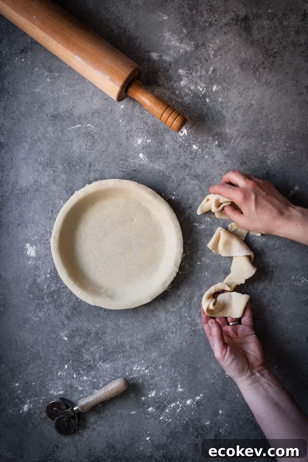 top view of hands trimming pie dough from pie tin with rolling pin lying nearby while making a rhubarb apple pie with ginger and lemongrass