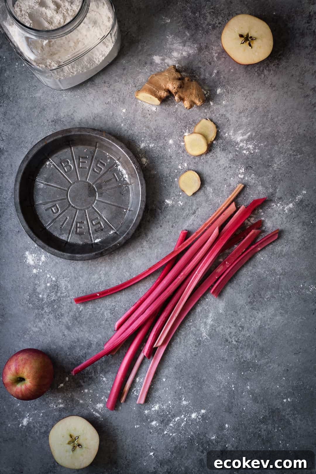 grey surface with metal pie tin and a glass jar of flour along with apples, rhubarb and ginger for rhubarb apple pie with ginger and lemongrass