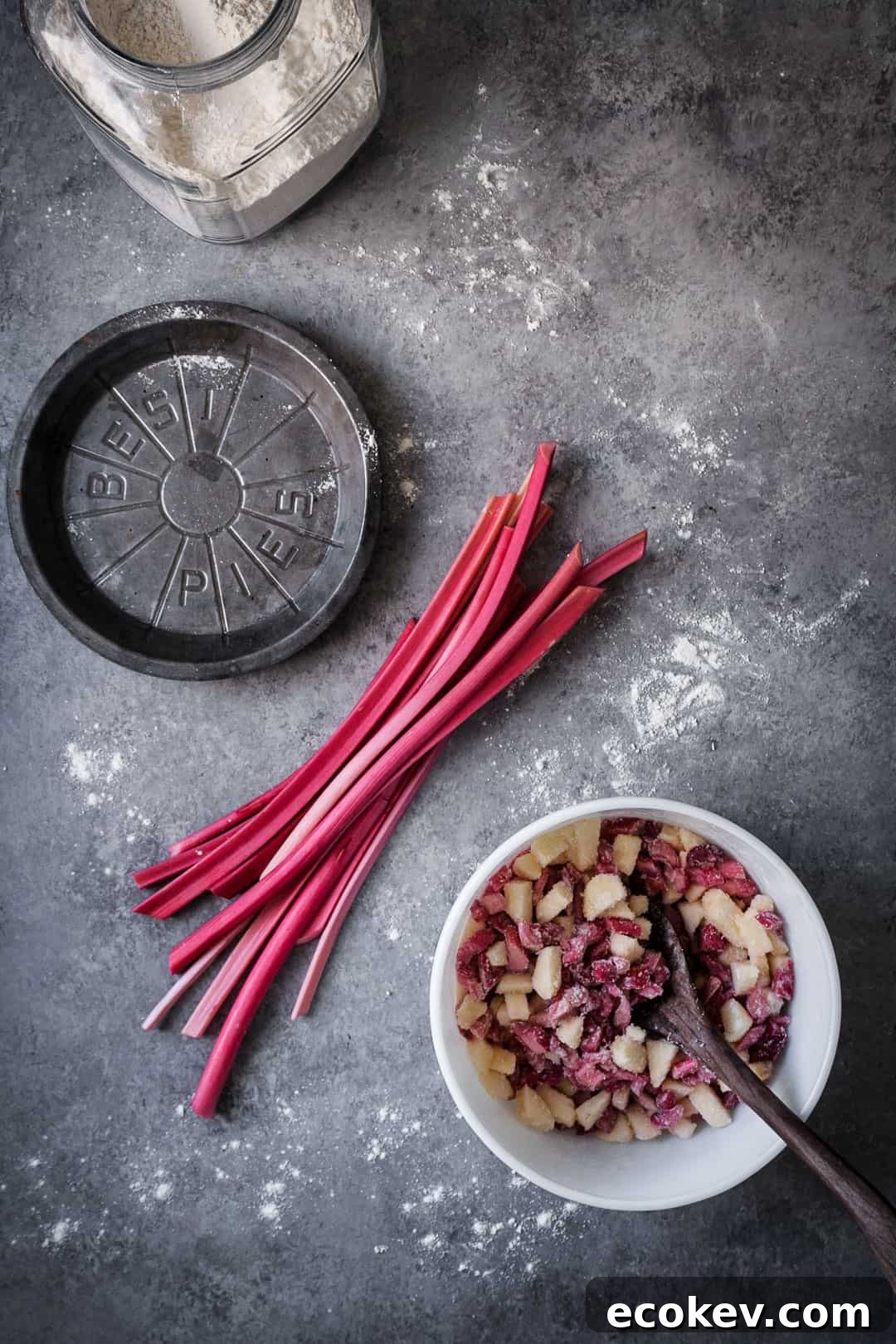 making a rhubarb apple pie with ginger and lemongrass: top view of rhubarb stalks on grey surface with metal pie tin and container of flour nearby along with white bowl filled with cut up apples and rhubarb
